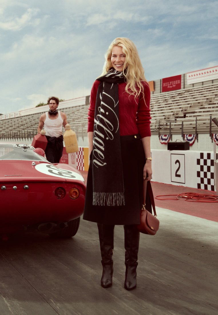 Suéter rojo, falda negra, bufanda larga negra con letras blancas, botas negras hasta la rodilla y un bolso marrón, al lado de un coche deportivo rojo.