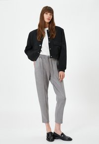 Mujer vestida con una chaqueta negra, camisa blanca, pantalones de tiro ajustable grises y mocasines negros, de pie contra un fondo blanco liso.