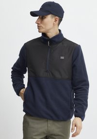 Pullover in pile navy con pannello in nylon nero sul petto, con zip al collo e tasche laterali. Presenta un patch con logo sul petto sinistro e polsini elasticizzati.