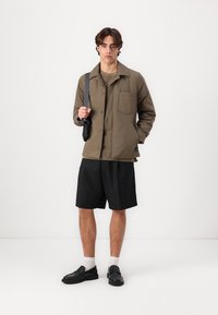 Olive green, button-up jacket with a single chest pocket; paired with black shorts, white socks, and black shoes. Smooth fabric, relaxed fit.
