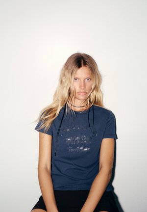 Jeune femme aux longs cheveux blonds portant un t-shirt bleu foncé avec un texte scintillant et un short noir sur un fond clair uni.