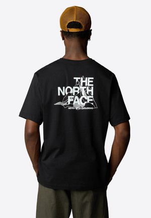Mann trägt ein schwarzes T-Shirt mit weißem "The North Face"-Schriftzug und Berggrafik auf dem Rücken, kombiniert mit olivgrüner Hose und brauner Mütze.