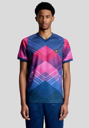 Giovane uomo che indossa una camicia a maniche corte con motivo geometrico in tonalità blu e rosa, in piedi davanti a uno sfondo grigio.