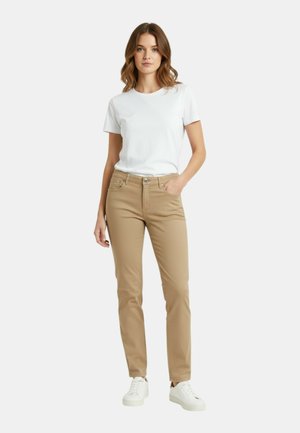 Donna in piedi che indossa una t-shirt bianca semplice, pantaloni beige aderenti e sneakers bianche, con una mano in tasca su uno sfondo neutro.