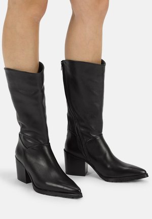 Schwarze Lederstiefel bis zur Mitte der Wade mit spitzen Zehen, Blockabsätzen und seitlichen Reißverschlüssen, getragen an nackten Beinen vor einem schlichten Hintergrund.