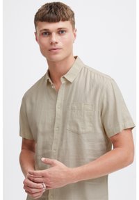 Kortærmet knap-op skjorte i beige linned med en brystlomme, ensartet tekstur og standard krave. Modellen poserer med hænderne foldet.