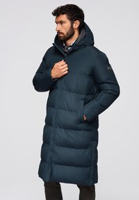 Marineblauwe lange pufferjas met een capuchon, voorzien van gestikte segmenten, voor ritsluiting en zijzakken. Gladde textuur met een matte afwerking.