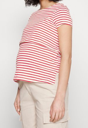Zwangere persoon draagt een rood-wit gestreept T-shirt met korte mouwen en beige cargobroek, staand tegen een effen achtergrond.