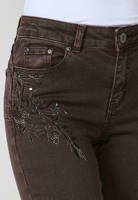 Jeans de mezclilla marrones con intrincados bordados con cuentas y motivos florales en la parte delantera del bolsillo izquierdo. Diseño estándar de cinco bolsillos.