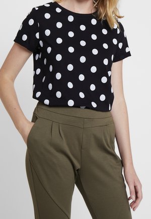 Femme portant une chemise noire à manches courtes à pois blancs rentrée dans un pantalon taille haute vert olive, une main dans la poche.