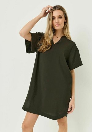 Vrouw met lang blond haar die een losse, korte mouw, donkergroene jurk draagt, poserend met één hand die haar haar aanraakt tegen een effen lichte achtergrond.