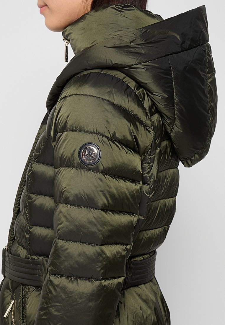 Person in olivgrünem steppbarem Pufferjacke mit schwarzer Kapuze und schwarzem Gürtel, Seitenansicht, die den Arm-Patch und die Reißverschlussdetails zeigt.