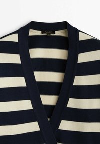 Cardigan tricoté à rayures horizontales bleu marine et blanc, avec un profond col en V et une bordure bleu marine foncé le long de l'encolure et des épaules.