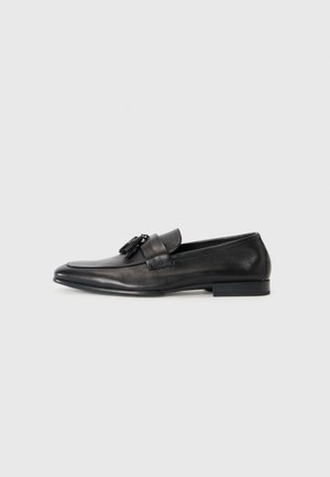 Mocassin en cuir noir avec pampilles, à talon bas et bout rond, profil latéral sur fond clair.