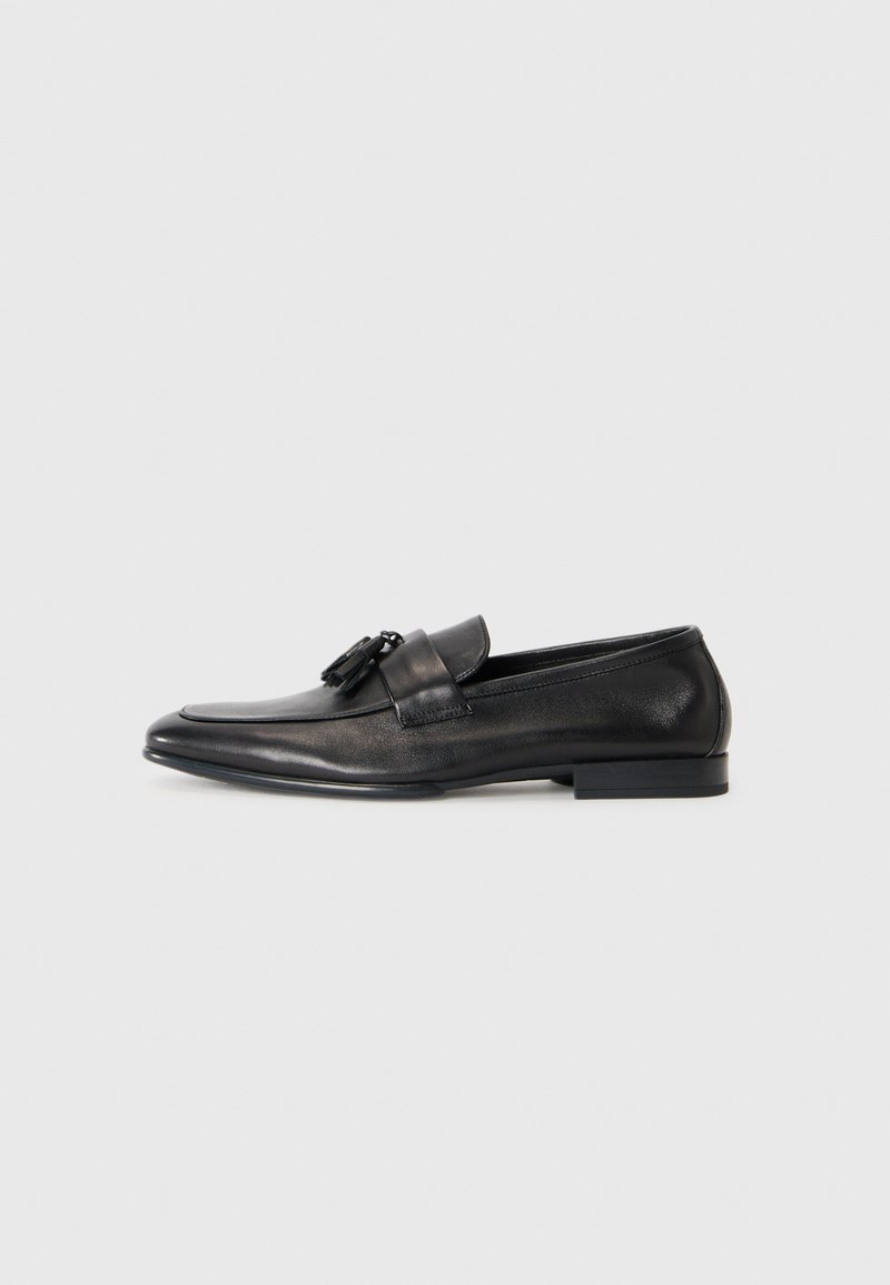 Mocassin en cuir noir avec pampilles, à talon bas et bout rond, profil latéral sur fond clair.