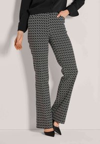 Vrouw in zwarte en witte geometrisch patroon wijde broek, zwarte puntige schoenen en een zwart long-sleeve top, met hand in de zak.