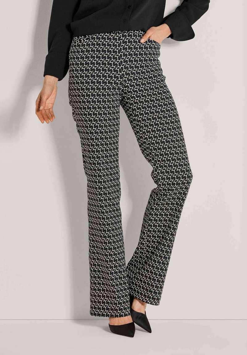 Vrouw in zwarte en witte geometrisch patroon wijde broek, zwarte puntige schoenen en een zwart long-sleeve top, met hand in de zak.