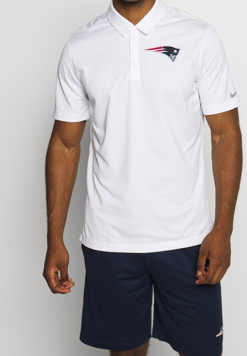 Homme portant un polo blanc avec le logo des New England Patriots et un short bleu marine, debout devant un fond uni.