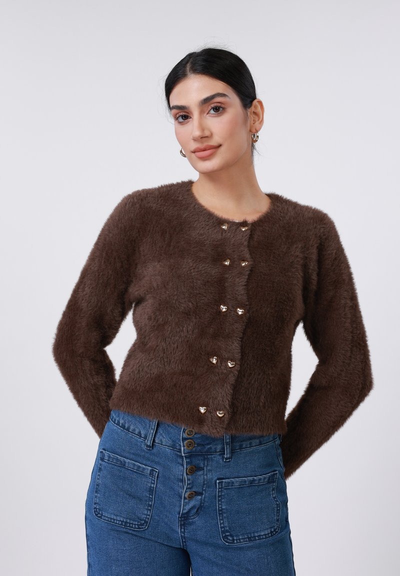 Pruun karvane kardigan südamekujude kuldsete nööpidega, ümmarguse kaelusega ja pikkade varrukatega, kombineeritud kõrge vöökohaga siniste teksapükstega.