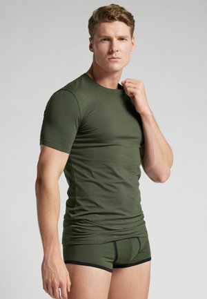 Mann mit kurzen Haaren, der ein eng anliegendes olivgrünes T-Shirt und passende Boxershorts trägt, posiert vor einem schlichten hellen Hintergrund.