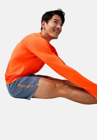 Orange långärmad träningströja med texturerad tyg, tillsammans med grå shorts med ordet "BROOKS" i orange. Modell som sträcker ut sig.