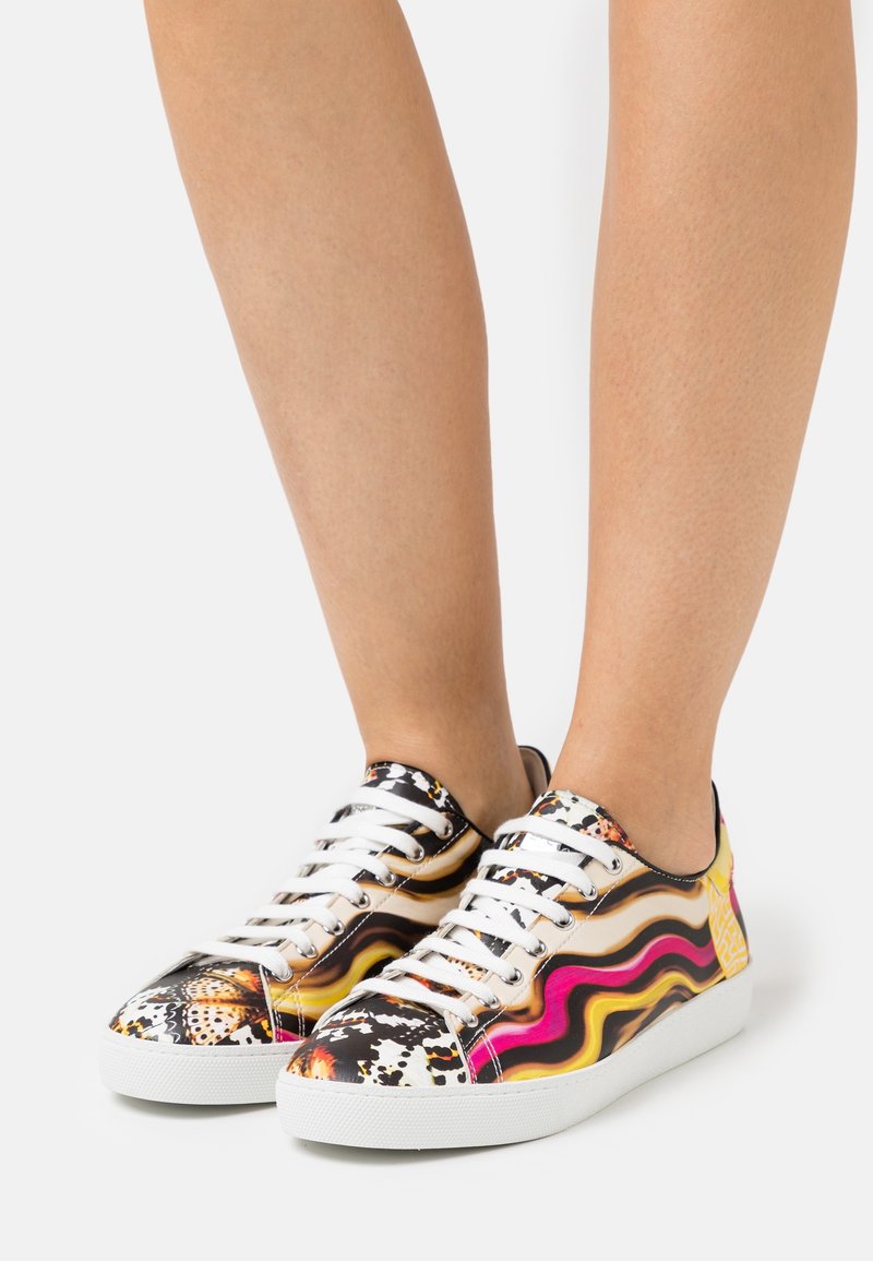 Jambes portant des baskets basses avec des lacets blancs décorés de motifs ondulés colorés en noir, jaune, rose et orange.