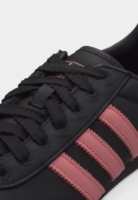 Μαύρο αθλητικό παπούτσι με τρεις ροζ ρίγες στο πλάι και μαύρα κορδόνια, που δείχνει το μπροστινό και πλαϊνό πάνω μέρος με λεπτομέρειες.