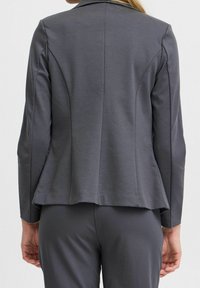Szara dopasowana marynarka z baskinką, usztywnionymi ramionami i długimi rękawami. Wykonana z gładkiej tkaniny, posiada gładki szew z tyłu i nie ma kieszeni.