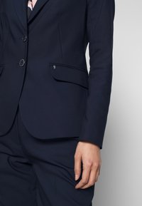 Buste et bras d'une femme portant un blazer bleu marine foncé sur mesure et un pantalon assorti, avec une chemise à motif visible au niveau du col.
