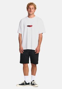 Hvid bomulds t-shirt med rund halsudskæring, korte ærmer, og et rødt og sort logo i midten. Båret sammen med sorte shorts og sorte sneakers.