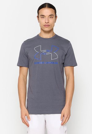 Homme debout, face à l'avant, portant un t-shirt gris Under Armour et un short blanc sur un fond blanc uni.