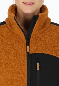Giacca in fleece marrone con colletto alto e accenti neri. Presenta una zip frontale e tasche laterali con zip, complete di piccoli tag del marchio.
