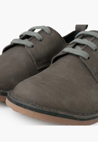 Scarpe in pelle marrone con punta arrotondata, lacci in tessuto grigio e suola in gomma beige. Texture opaca con dettagli di cucitura sottili.