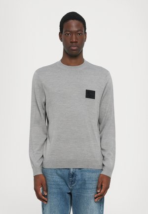 Gray long-sleeve sweater made of knit fabric. Features a textured black patch on the chest. Round neckline and ribbed cuffs. Worn with blue jeans.