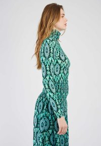 Robe à motifs verts avec un col montant et des manches longues, dotée d'une taille froncée et d'une silhouette fluide. Les tissus ont une texture lisse.