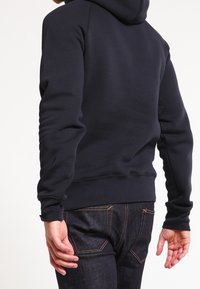 Felpa blu navy con una vestibilità rilassata, dotata di cappuccio e polsini a costine, abbinata a jeans in denim scuro con dettagli di cucitura a contrasto.