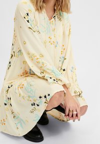 Květinové šaty ze světlého žlutého materiálu, s dlouhými rukávy, výstřihem do V a jemnou výšivkou v modré, oranžové a černé akcenty.