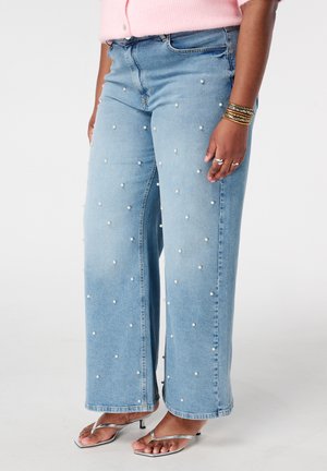 Lichtblauwe wijduitlopende spijkerbroek met verspreide witte parels, gedragen met zilveren sandalen met hak en gouden armbanden aan één pols.