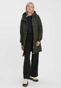 Cappotto verde trapuntato con cappuccio, dotato di bottoni a pressione, maniche lunghe e taglio dritto. Abbinato a pantaloni neri a gamba larga e stivali beige.