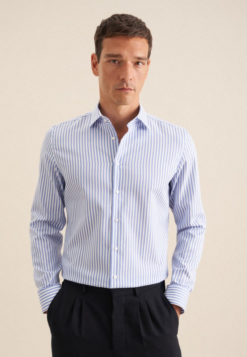 Homme portant une chemise boutonnée à rayures verticales bleu clair et blanc avec un pantalon noir, debout les mains dans les poches devant un fond uni.