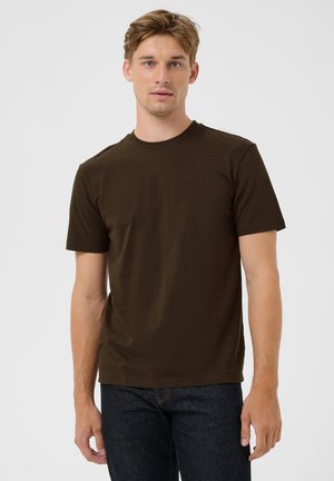 Joven con cabello castaño claro que lleva una camiseta de manga corta marrón oscuro lisa y vaqueros azul oscuro, de pie frente a un fondo claro.
