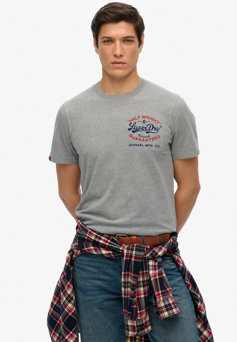 T-shirt gris à manches courtes avec un logo en bleu marine et rouge. Présente une chemise à carreaux nouée à la taille et un jean en denim bleu. Design simple.