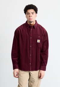 Burgundy kordfløjl skjorte med knapper, fra brystlommen og logo-patch, har en afslappet pasform og lange ærmer. Lyse beige bukser.