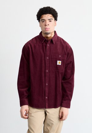 Burgundin värinen corduroy-nappipaita, jossa on rintatasku ja logopatch, rento istuvuus ja pitkät hihat. Vaaleanbeiget housut.