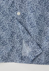 Chemise à motifs bleus et blancs avec des motifs tourbillonnants, fermeture par boutons, texture de tissu lisse avec une fente sur le côté et des poignets repliés.