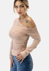 Långärmad off-shoulder topp i ljusrosa tyg med rynkad textur. Matchad med blå jeans och accentuerad av en hjärt-tatuering.