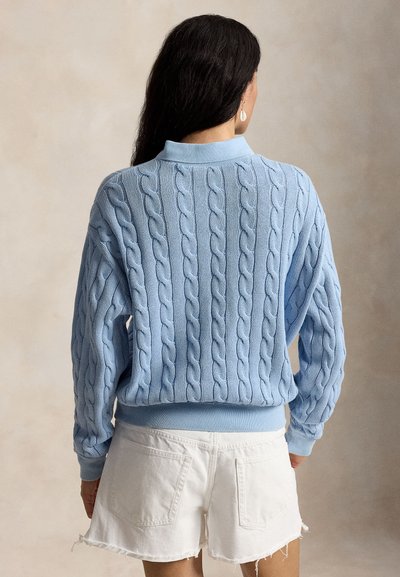 Kobieta z długimi ciemnymi włosami, ubrana w jasno-niebieski sweter w warkoczowy splot i białe szorty jeansowe, stojąca tyłem do kamery.