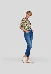 Gemustertes Langarmoberteil in Grün-, Braun- und Cremetönen, kombiniert mit enganliegenden blauen Jeans und weißen Sneakers mit sandfarbenen Akzenten.