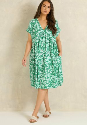 Frau steht und trägt ein grünes knielanges Kleid mit weißen Blattmustern und weiße Slip-On-Sandalen vor beigem Hintergrund.