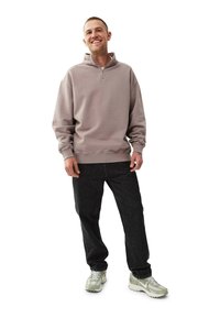 Vaaleanharmaa vetoketjullinen huppari, jossa on korkea kaulus, yhdistettynä tummiin farkkuihin ja vaaleisiin lenkkareihin. Kangas näyttää pehmeältä ja sillä on rento istuvuus.